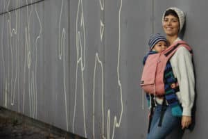 Frau mit rosaner Didymos babytrage (didysnap) lehnt an der Wand
